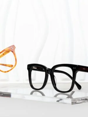 Elegant glass frames in black and tortoise colors showcased on a clear acrylic stand against a wavy backdrop.